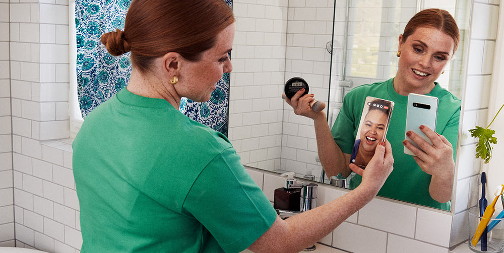 Tjej som ser in i spegel med grön tröja och ser glad ut, facetimear med en vän